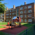Property for sale by Auction A playground with a climbing frame and slide sits on a red rubber surface, enclosed by a black fence at 21 Glazebrook Close in Dulwich, London. Behind it is a four-story brick apartment building with multiple windows. Trees provide shade to the left, and there is a clear blue sky above. Presented by Palace Auctions