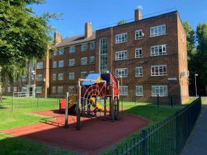 Property for sale by Auction A playground with a climbing frame and slide sits on a red rubber surface, enclosed by a black fence at 21 Glazebrook Close in Dulwich, London. Behind it is a four-story brick apartment building with multiple windows. Trees provide shade to the left, and there is a clear blue sky above. Presented by Palace Auctions