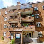 Property for sale by Auction A four-story brick apartment building with balconies, featuring plants and household items, stands proudly at 21 Glazebrook Close, Dulwich. The ground floor has an entrance with a notice board and a curved, stone planter. The sky is bright and partly cloudy. The surroundings are grassy with a road to the left. Presented by Palace Auctions