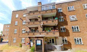 Property for sale by Auction A four-story brick apartment building with balconies, featuring plants and household items, stands proudly at 21 Glazebrook Close, Dulwich. The ground floor has an entrance with a notice board and a curved, stone planter. The sky is bright and partly cloudy. The surroundings are grassy with a road to the left. Presented by Palace Auctions