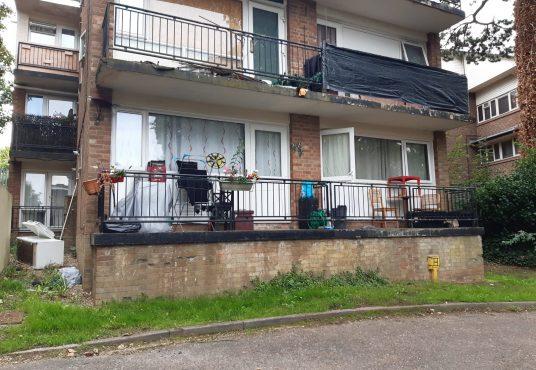 Property for sale by Auction A photo of a two-story, brick apartment building at 37 Farley Lodge, Bedfordshire, with balconies on both levels. The upper balcony has black tarpaulin covering its railing. The lower balcony in Luton is furnished with chairs, potted plants, and small decorations. The grass in front of the building is untidy. Presented by Palace Auctions