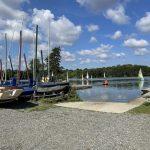 Property for sale by Auction A lakeside view with multiple sailboats docked on the shore and some out on the water. The sky is bright blue with scattered fluffy clouds. A gravel area leads down to a small ramp, providing access to the lake. Trees line the far shore in the background near plots of land at 435 Chevening Road, Sevenoaks. Presented by Palace Auctions