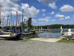 Property for sale by Auction A lakeside view with multiple sailboats docked on the shore and some out on the water. The sky is bright blue with scattered fluffy clouds. A gravel area leads down to a small ramp, providing access to the lake. Trees line the far shore in the background near plots of land at 435 Chevening Road, Sevenoaks. Presented by Palace Auctions