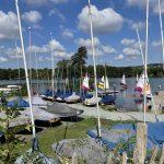 Property for sale by Auction A scenic view of a lake on a sunny day near Sevenoaks, TN13 2SA, with blue skies and scattered clouds. Numerous sailboats with colorful sails are anchored or sailing on the water, and several masts are prominently visible. The shoreline is lined with green grass and plots of land at 435 Chevening Road in the background. Presented by Palace Auctions