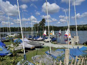 Property for sale by Auction A scenic view of a lake on a sunny day near Sevenoaks, TN13 2SA, with blue skies and scattered clouds. Numerous sailboats with colorful sails are anchored or sailing on the water, and several masts are prominently visible. The shoreline is lined with green grass and plots of land at 435 Chevening Road in the background. Presented by Palace Auctions