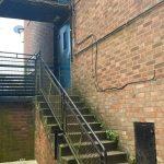 Property for sale by Auction A narrow outdoor staircase with a metal railing leads up to a blue door in the brick building at 37 Farley. The surrounding area looks weathered and slightly overgrown with moss and plants. An abandoned piece of fabric lies at the bottom of the stairs. Presented by Palace Auctions