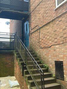 Property for sale by Auction A narrow outdoor staircase with a metal railing leads up to a blue door in the brick building at 37 Farley. The surrounding area looks weathered and slightly overgrown with moss and plants. An abandoned piece of fabric lies at the bottom of the stairs. Presented by Palace Auctions