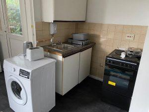 Property for sale by Auction A small kitchen at 37 Farley features a washing machine, toaster, and sink on the left. A stove with an oven and various items on top is on the right. Cabinets above and below the sink and a door with a window next to the washing machine are visible. The floor is dark-colored. Presented by Palace Auctions