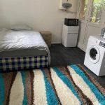 Property for sale by Auction A small room at 37 Farley with a single bed, a nightstand, and a colorful striped rug on a dark floor. There is a white washing machine, mini fridge, microwave, and kettle beneath a wall-mounted boiler. A door leads outside. Presented by Palace Auctions