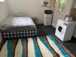 Property for sale by Auction A small room at 37 Farley with a single bed, a nightstand, and a colorful striped rug on a dark floor. There is a white washing machine, mini fridge, microwave, and kettle beneath a wall-mounted boiler. A door leads outside. Presented by Palace Auctions