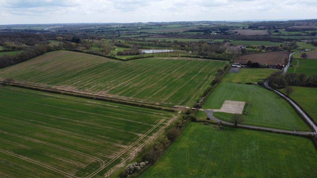 Property for sale by Auction Aerial view of expansive farmland, showcasing large green and brown fields divided by hedgerows at Epping Farm. In the background, trees, a small lake, and scattered buildings are visible. The landscape extends towards a horizon under a partly cloudy sky, captured beautifully by drone. Presented by Palace Auctions
