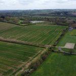 Property for sale by Auction Aerial view of expansive farmland, showcasing large green and brown fields divided by hedgerows at Epping Farm. In the background, trees, a small lake, and scattered buildings are visible. The landscape extends towards a horizon under a partly cloudy sky, captured beautifully by drone. Presented by Palace Auctions