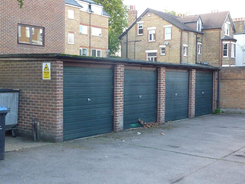 Property for sale by Auction Four closed garage doors made of dark green metal are situated within a brick structure in Beech Court. The garages are part of a residential complex with surrounding brick buildings. A "No Parking" sign is visible on the left side, along with trash bins and scattered debris. Presented by Palace Auctions