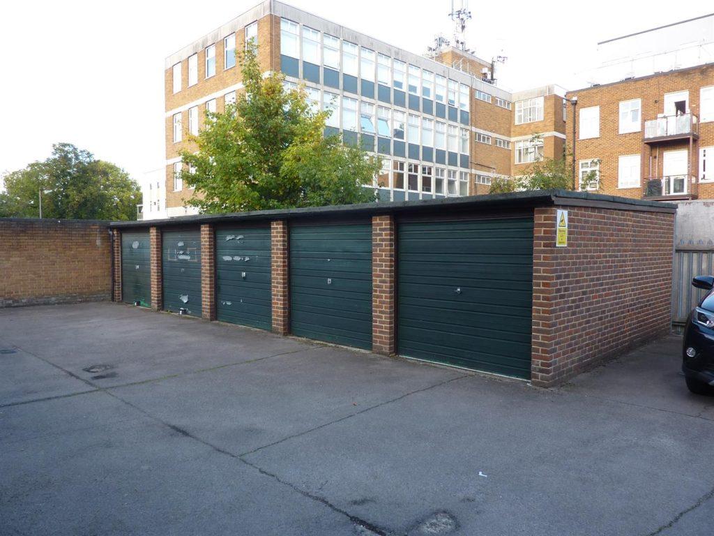 Property for sale by Auction A row of four green garage doors on a brick structure, forming the outdoor garage unit at Beech Court. The background features a multi-story building with numerous windows and a tree. The ground is paved, with part of a car visible on the right side of the image. Presented by Palace Auctions