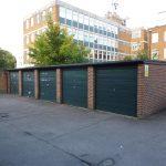 Property for sale by Auction A row of four green garage doors on a brick structure, forming the outdoor garage unit at Beech Court. The background features a multi-story building with numerous windows and a tree. The ground is paved, with part of a car visible on the right side of the image. Presented by Palace Auctions