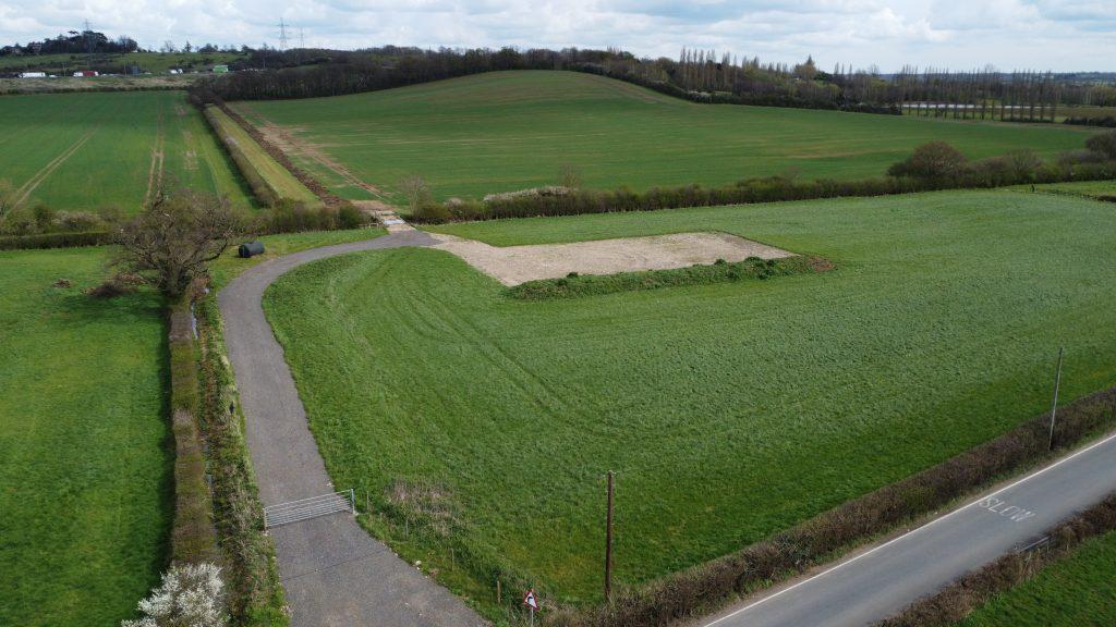 Property for sale by Auction Aerial view of a vast green field at Epping Farm, with a rectangular dirt patch in the center. A narrow paved road runs along the bottom edge, intersecting another smaller path leading towards the dirt area. Trees and hedges surround the field, with gently rolling hills in the background. Presented by Palace Auctions