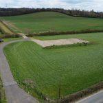 Property for sale by Auction Aerial view of a vast green field at Epping Farm, with a rectangular dirt patch in the center. A narrow paved road runs along the bottom edge, intersecting another smaller path leading towards the dirt area. Trees and hedges surround the field, with gently rolling hills in the background. Presented by Palace Auctions