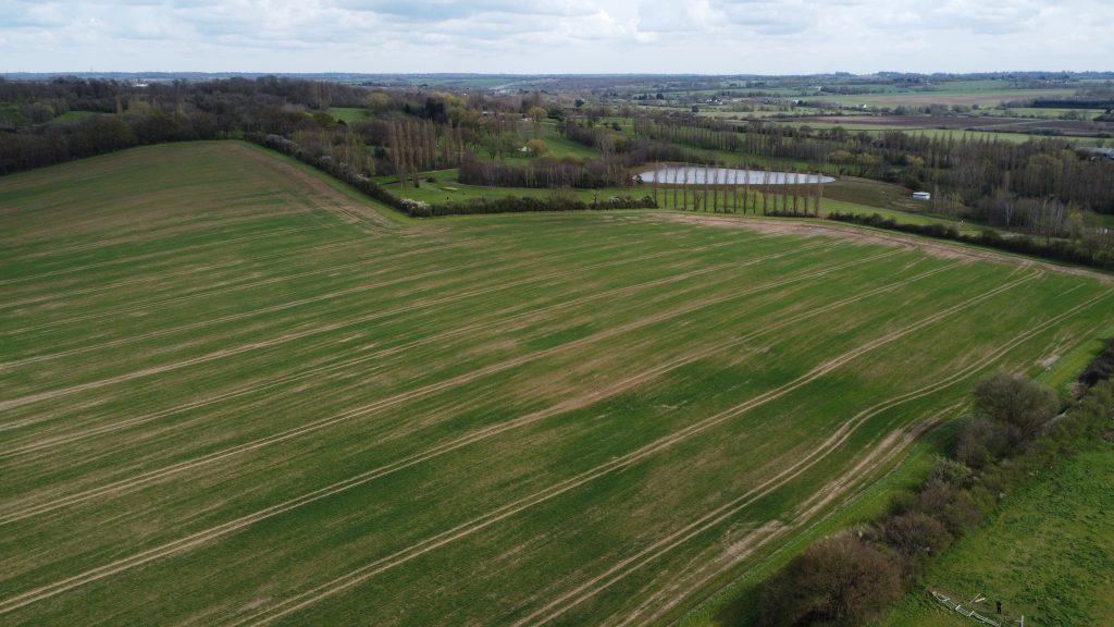Property for sale by Auction Aerial views taken by drone reveal Epping Farm's expansive, green fields with rows of crops. The landscape showcases gently rolling hills, patches of trees, and a small lake in the distance under a cloudy sky with hints of blue peeking through. Presented by Palace Auctions