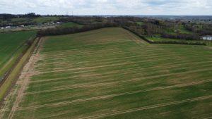 Property for sale by Auction Aerial view of the vast green fields at Epping Farm, with several dirt tracks running through them, surrounded by rows of trees and bordered by more greenery. The sky is cloudy, and hills and distant structures are visible on the horizon. Presented by Palace Auctions