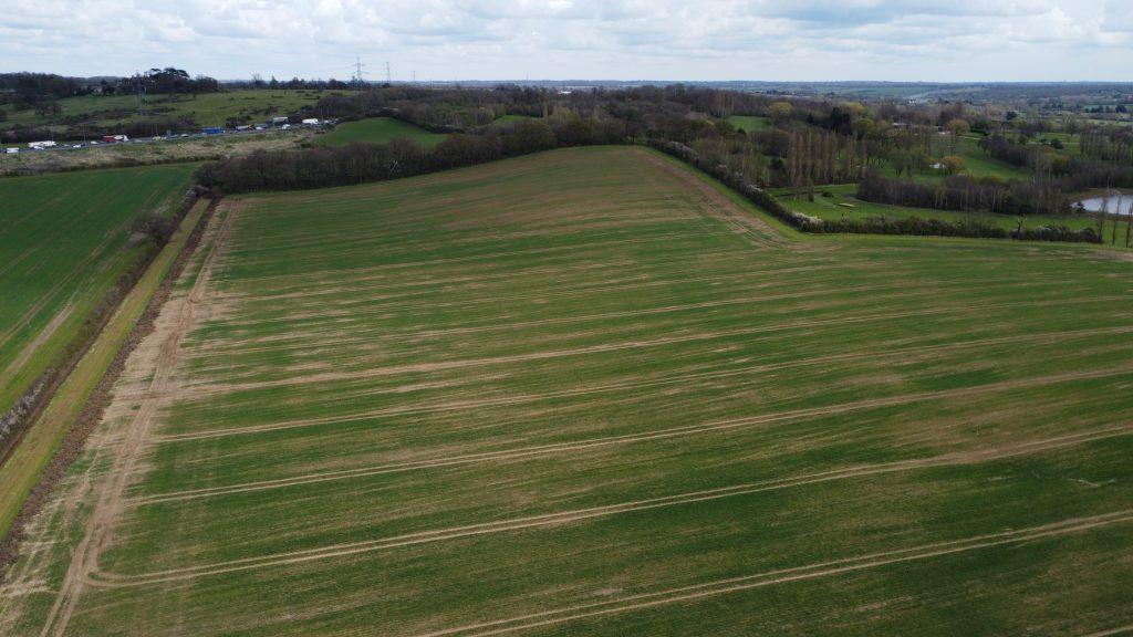Property for sale by Auction Aerial view of a vast green field with subtle lines of brown soil, bordered by trees and hedges on two sides. In the distance at Epping Farm, there are more fields, a cluster of buildings, and scattered trees under a cloudy sky; drone views capture this serene landscape. Presented by Palace Auctions