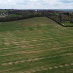Property for sale by Auction Aerial view of a vast green field with subtle lines of brown soil, bordered by trees and hedges on two sides. In the distance at Epping Farm, there are more fields, a cluster of buildings, and scattered trees under a cloudy sky; drone views capture this serene landscape. Presented by Palace Auctions