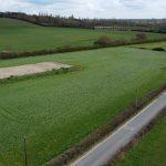 Property for sale by Auction An aerial view taken by drone shows a rural landscape with green fields at Epping Farm, along with a small sandy area in one field and a narrow country road curving alongside the fields. Trees and hedges divide the fields, while distant woods and scattered buildings are visible on the horizon under a cloudy sky. Presented by Palace Auctions