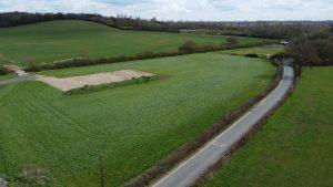 Property for sale by Auction An aerial view taken by drone shows a rural landscape with green fields at Epping Farm, along with a small sandy area in one field and a narrow country road curving alongside the fields. Trees and hedges divide the fields, while distant woods and scattered buildings are visible on the horizon under a cloudy sky. Presented by Palace Auctions