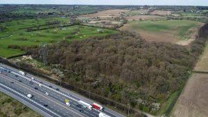 Property for sale by Auction Aerial views taken by drone reveal a highway with vehicles next to a dense wooded area bordered by expansive fields, including land at Epping Farm. The landscape features patches of green farmland and clusters of trees extending towards distant hills under a clear sky with scattered clouds. Presented by Palace Auctions