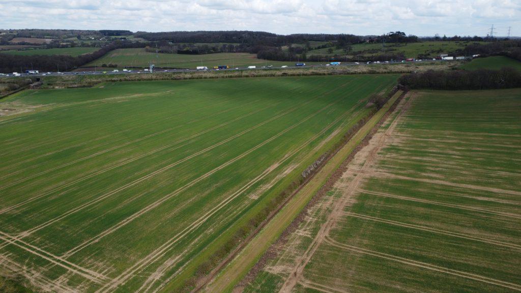 Property for sale by Auction Aerial view of a large green field at Epping Farm, divided by a dirt path. The field is surrounded by trees with rolling hills in the background. A highway with moving vehicles can be seen in the distance under a cloudy sky, showcasing captivating drone views taken by Palace Auctions. Presented by Palace Auctions
