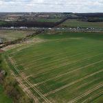 Property for sale by Auction Aerial view of Epping Farm reveals a large green field with some brown patches, surrounded by trees and neighboring fields. To the left, there are a few small buildings and parked cars. A highway with vehicles cuts across the landscape in the background, under a partly cloudy sky. Presented by Palace Auctions