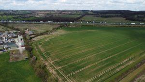 Property for sale by Auction Aerial view of Epping Farm reveals a large green field with some brown patches, surrounded by trees and neighboring fields. To the left, there are a few small buildings and parked cars. A highway with vehicles cuts across the landscape in the background, under a partly cloudy sky. Presented by Palace Auctions