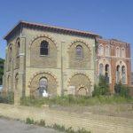 Property for sale by Auction An old, abandoned brick building with arched windows, partially deteriorated, and overgrown vines covering some sections. Standing on an unkempt lot with scattered wild plants under a clear blue sky, it evokes memories of Sheerness before the Water Tower development transformed the area. Presented by Palace Auctions