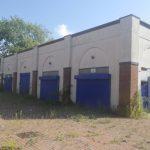 Property for sale by Auction A brick building with blue shutters on each of its three visible sections, surrounded by overgrown weeds and tall grass. The building appears to be abandoned, under a partly cloudy sky with a tree in the background. Nearby houses of The Water Tower development in Sheerness are also partially visible. Presented by Palace Auctions