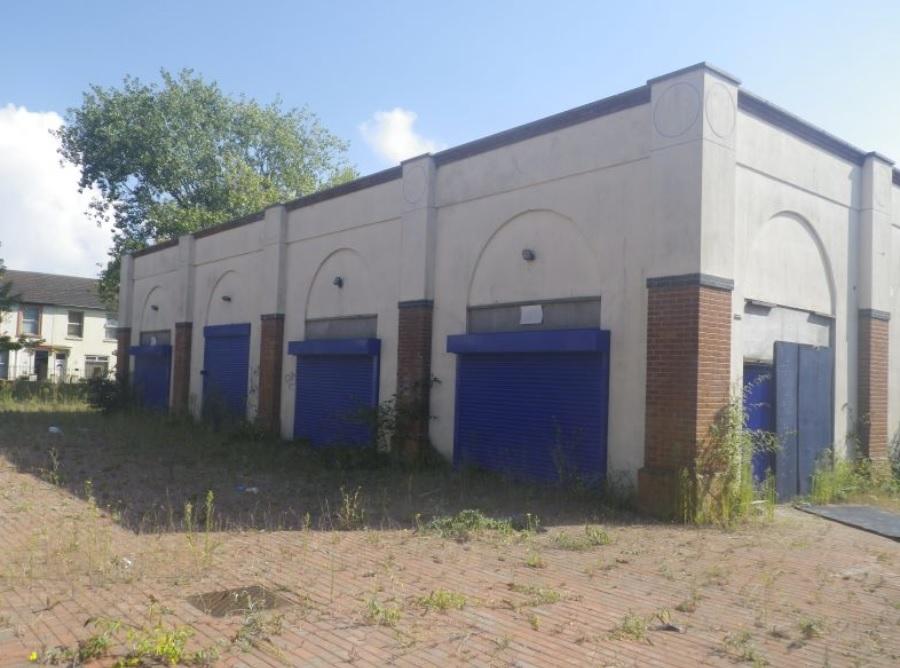 Property for sale by Auction A brick building with blue shutters on each of its three visible sections, surrounded by overgrown weeds and tall grass. The building appears to be abandoned, under a partly cloudy sky with a tree in the background. Nearby houses of The Water Tower development in Sheerness are also partially visible. Presented by Palace Auctions