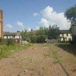 Property for sale by Auction A vacant, overgrown lot with patches of weeds and greenery. To the left, an old, partly dilapidated brick building, and to the right, a row of garages with blue doors. Residential houses and trees are visible in the background under a blue sky with scattered clouds near The Water Tower development in Sheerness. Presented by Palace Auctions