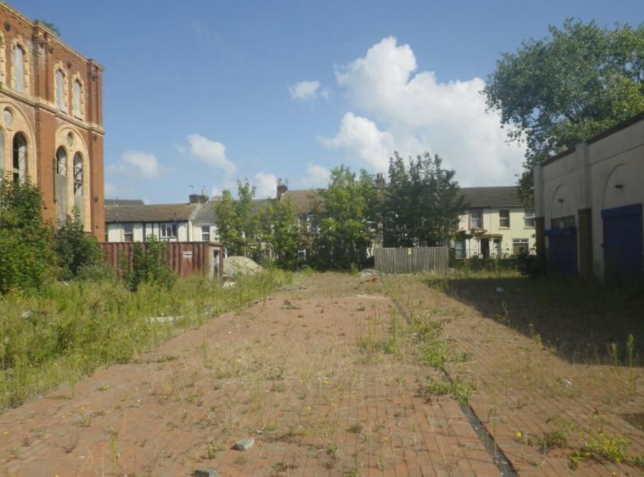 Property for sale by Auction A vacant, overgrown lot with patches of weeds and greenery. To the left, an old, partly dilapidated brick building, and to the right, a row of garages with blue doors. Residential houses and trees are visible in the background under a blue sky with scattered clouds near The Water Tower development in Sheerness. Presented by Palace Auctions