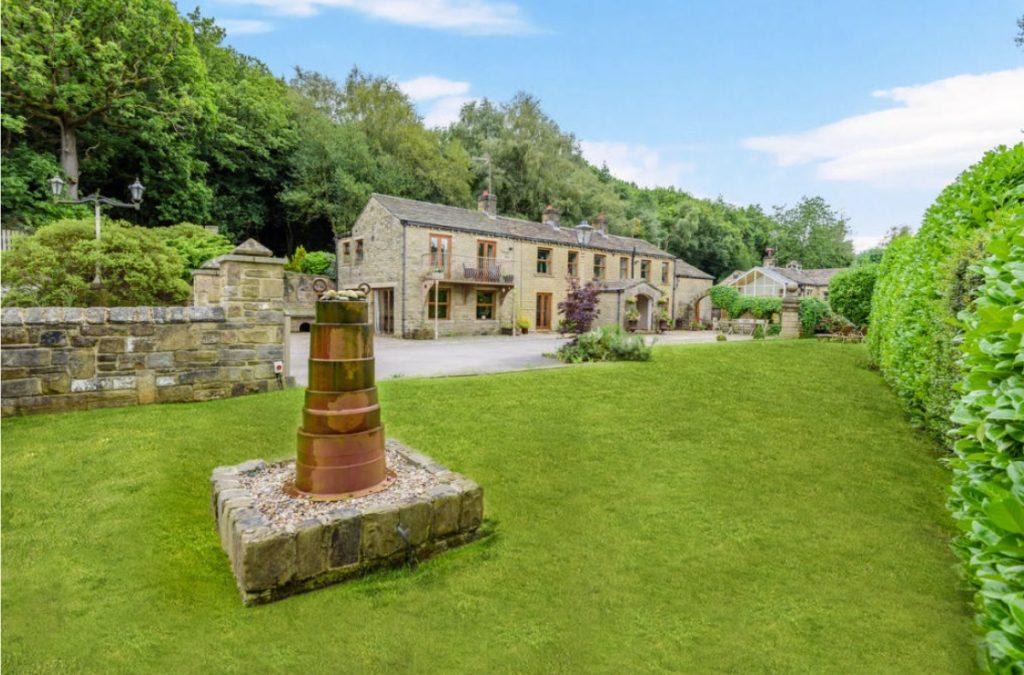 Property for sale by Auction A picturesque stone house with a manicured lawn and lush greenery on Bentley Road, Holmfirth. The house boasts a charming rustic appearance with multiple windows and a stone path leading up to it. In the foreground, there is a cylindrical stone structure on a stone base. Trees frame the background. HD9 4AP Presented by Palace Auctions