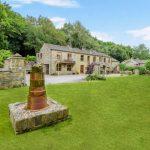 Property for sale by Auction A picturesque stone house with a manicured lawn and lush greenery on Bentley Road, Holmfirth. The house boasts a charming rustic appearance with multiple windows and a stone path leading up to it. In the foreground, there is a cylindrical stone structure on a stone base. Trees frame the background. HD9 4AP Presented by Palace Auctions