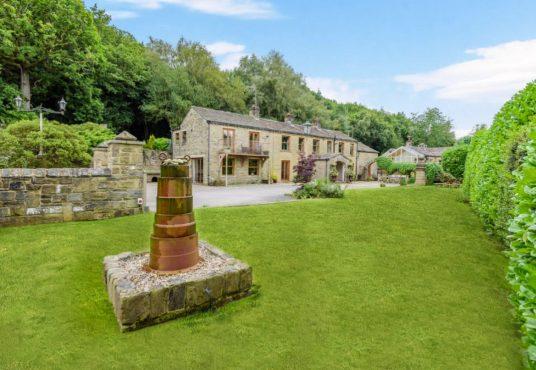 Property for sale by Auction A picturesque stone house with a manicured lawn and lush greenery on Bentley Road, Holmfirth. The house boasts a charming rustic appearance with multiple windows and a stone path leading up to it. In the foreground, there is a cylindrical stone structure on a stone base. Trees frame the background. HD9 4AP Presented by Palace Auctions