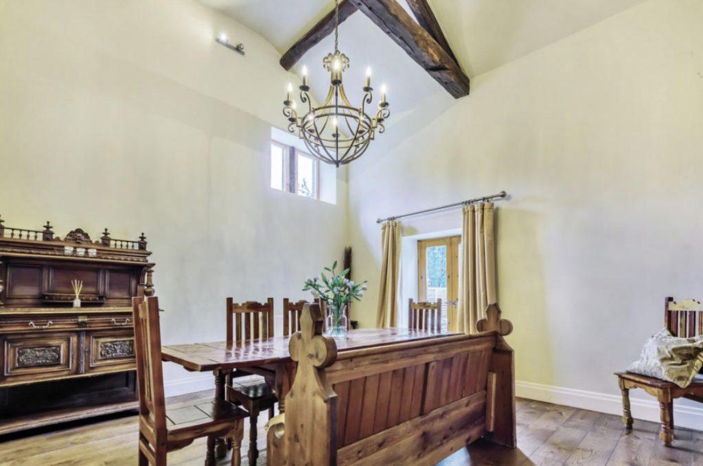 Property for sale by Auction A dining room on Bentley Road, Holmfirth, with high ceilings and exposed wooden beams features a wooden dining table with six matching chairs. A decorative chandelier hangs above the table, and a rustic wooden sideboard stands against the wall. A door with curtains leads outside. Presented by Palace Auctions
