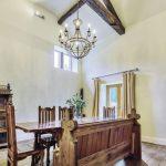 Property for sale by Auction A dining room on Bentley Road, Holmfirth, with high ceilings and exposed wooden beams features a wooden dining table with six matching chairs. A decorative chandelier hangs above the table, and a rustic wooden sideboard stands against the wall. A door with curtains leads outside. Presented by Palace Auctions