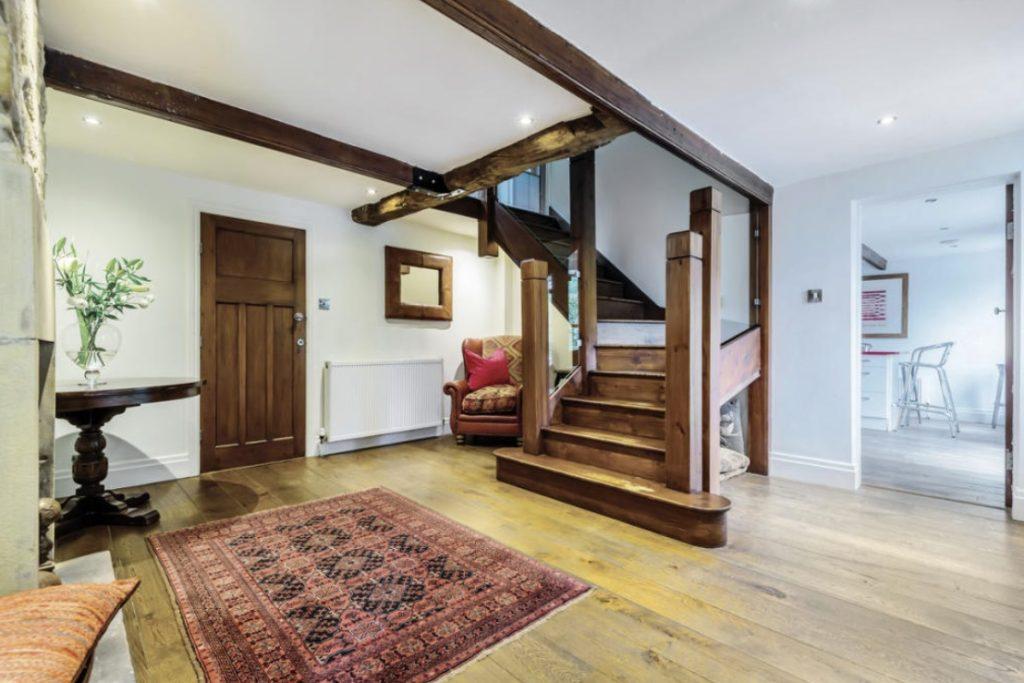 Property for sale by Auction A cozy hallway on Bent Ley Road, HOLMFIRTH, with wooden floors and a staircase featuring chunky wooden beams. There is a red patterned rug on the floor, a small round table with a vase of flowers to the left, and an armchair with cushions behind the stairs. A wooden door and a radiator are visible. Presented by Palace Auctions