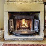 Property for sale by Auction A cozy living room on Bentley Road, Holmfirth, features a stone fireplace with a roaring fire. The fireplace has an open metal door and is set against a stone wall. Nearby, a wicker basket with firewood and a metal container are placed. Partial views of armchairs and a rug are visible in the background. Presented by Palace Auctions