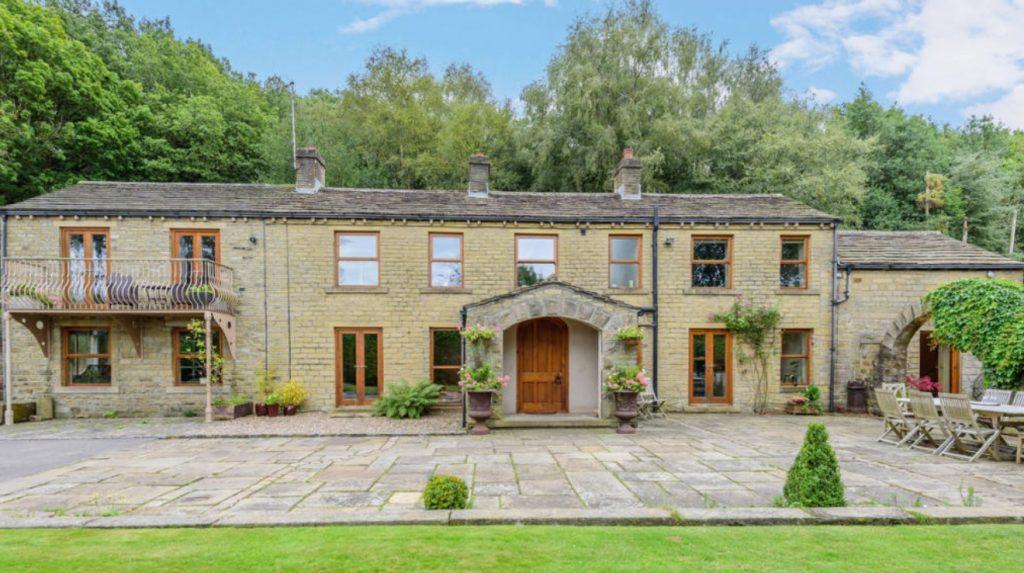 Property for sale by Auction A spacious two-story stone house on Bentley Road, HD9 4AP, Holmfirth features multiple windows and wooden doors surrounded by greenery. The front yard offers a paved stone patio with potted plants and outdoor seating. A balcony adorns the left side against a lush, tree-filled area under a blue sky. Presented by Palace Auctions