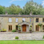 Property for sale by Auction A spacious two-story stone house on Bentley Road, HD9 4AP, Holmfirth features multiple windows and wooden doors surrounded by greenery. The front yard offers a paved stone patio with potted plants and outdoor seating. A balcony adorns the left side against a lush, tree-filled area under a blue sky. Presented by Palace Auctions