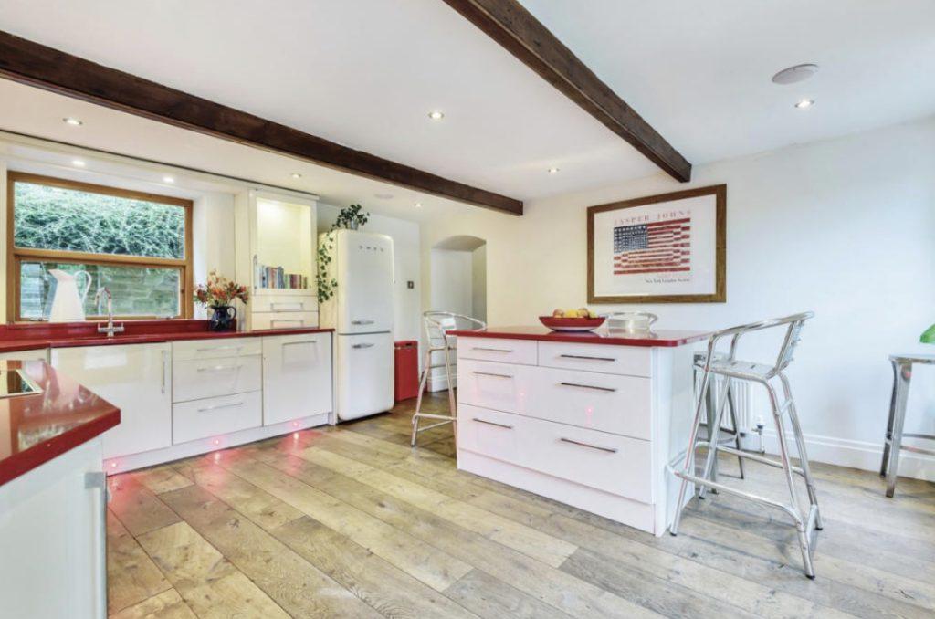 Property for sale by Auction A modern kitchen with white cabinets and red countertops in Holmfirth, HD9 4AP. An island with bar stools is centered. There's a vintage-style white refrigerator, a wooden floor, exposed wooden ceiling beams, a large window, and a framed American flag decor on the wall. Presented by Palace Auctions