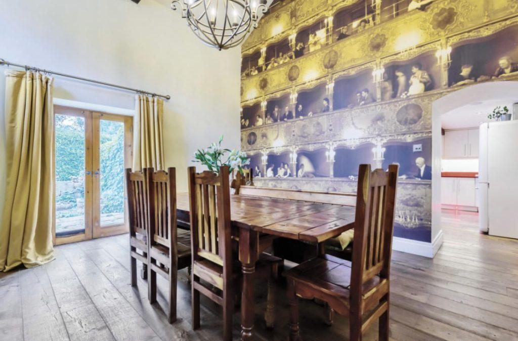 Property for sale by Auction A cozy dining room on Bentley Road, HD9 4AP, features a wooden table with six matching chairs. A chandelier hangs from the ceiling. One wall is adorned with a mural depicting a vintage theater audience. French doors with yellow curtains offer a view of greenery outside, typical of charming Holmfirth homes. Presented by Palace Auctions