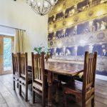 Property for sale by Auction A cozy dining room on Bentley Road, HD9 4AP, features a wooden table with six matching chairs. A chandelier hangs from the ceiling. One wall is adorned with a mural depicting a vintage theater audience. French doors with yellow curtains offer a view of greenery outside, typical of charming Holmfirth homes. Presented by Palace Auctions