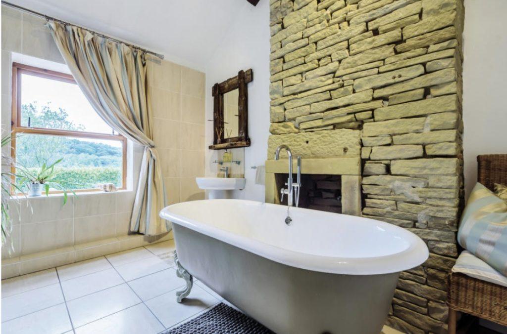 Property for sale by Auction A bathroom on Bentley Road, HD9 4AP, features a stone feature wall, a freestanding clawfoot bathtub, a wicker chair with cushions, and a window with light-colored curtains. The floor is tiled, and there is a small sink with a mirrored wooden frame on the wall above it. Presented by Palace Auctions