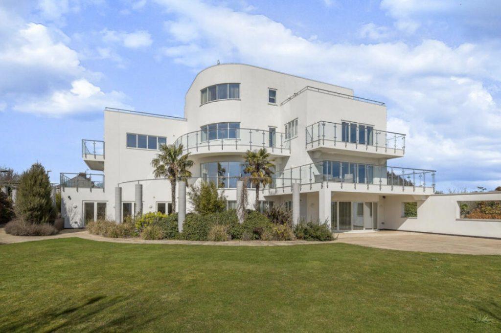 Property for sale by Auction A modern, multi-story white house with extensive glass panel balconies and large windows, set in a well-maintained garden on Dilks Street. The house features several rounded and angular architectural elements and is surrounded by lush green grass and a few palm trees. The sky is partly cloudy. Presented by Palace Auctions